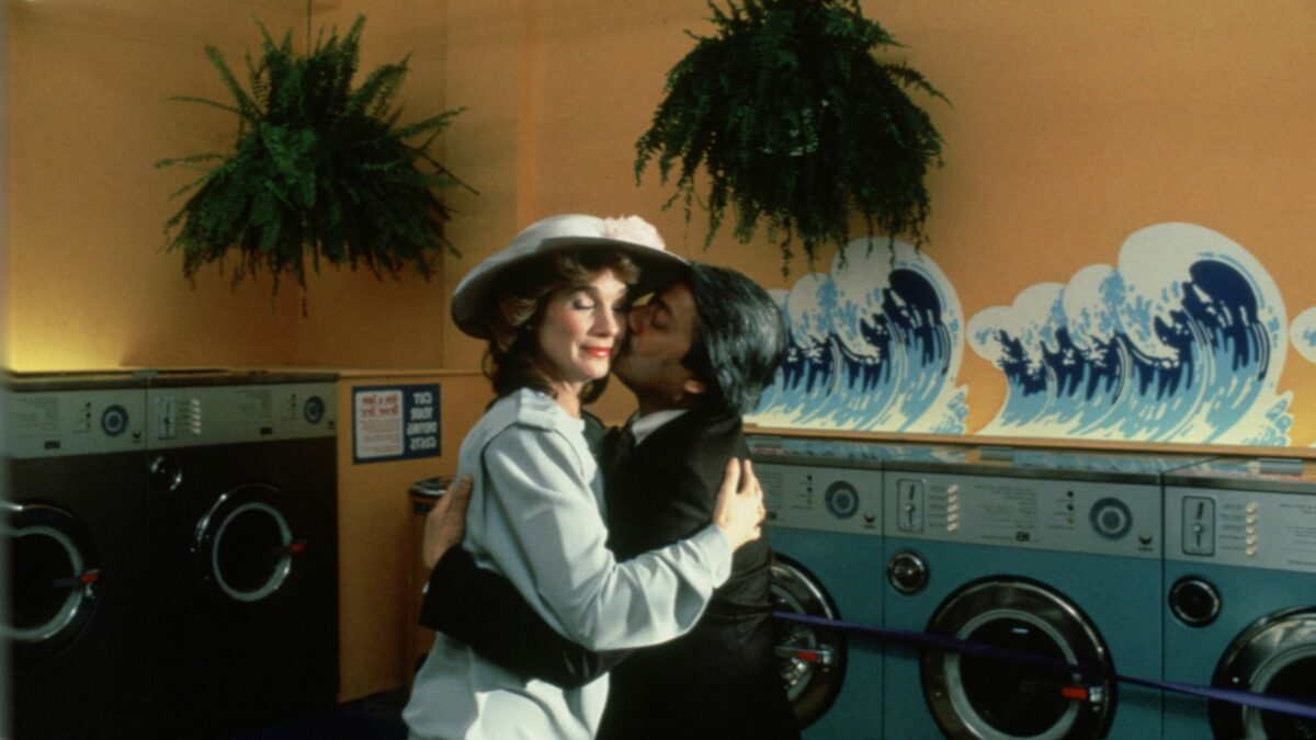 Mężczyzna obejmuje i całuje w policzek kobietę w kapeluszu. W tle stoją pralki przy malowidle przedstawiającym morskie fale.