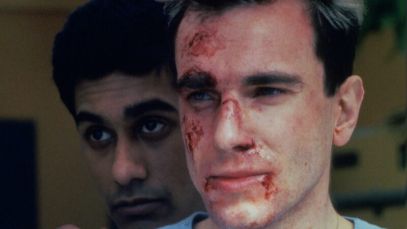 Zakrwawiona twarz młodego mężczyzny. Za nią znajduje się druga męska twarz. Obie patrzą w dal.