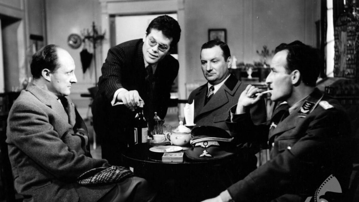 Grupa mężczyzn przy stole, jeden częstuje pozostałych alkoholem.