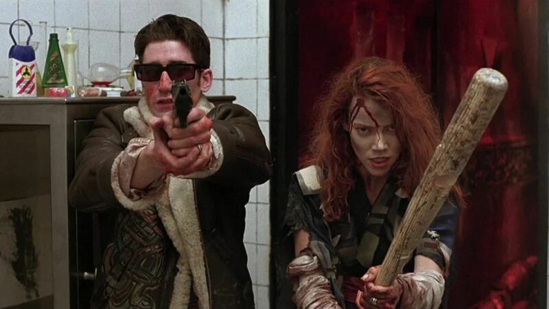 Mężczyzna trzymający pistolet, obok niego stoi kobieta z kijem bejsbolowym.