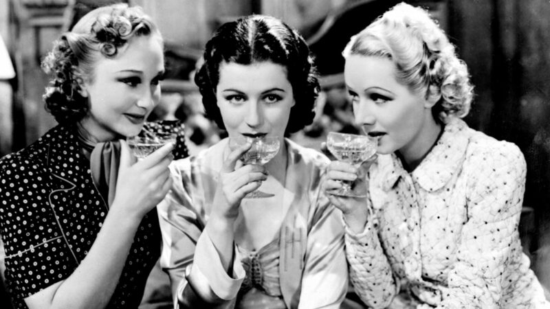 Trzy kobiety ubrane i uczesane w stylu lat trzydziestych piją drinki z szklanych kieliszków.