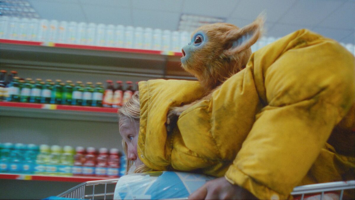 Dziewczynka biegnie z wózkiem sklepowym po alejkach marketu, na plecach ma dziwnego stwora.